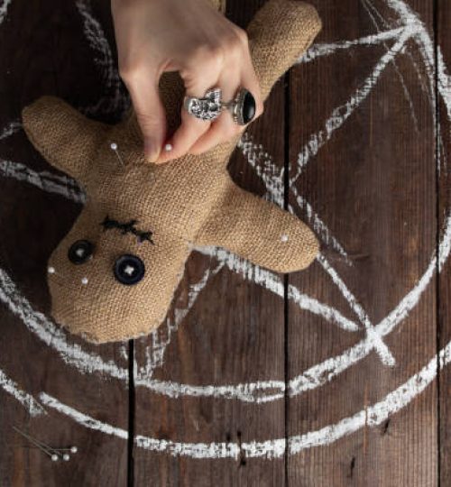 Voodoo doll on a wooden background with dramatic lighting. The concept of witchcraft and black art and the occult. Burlap doll on the background of a drawn star. Hand sticking pins into a doll. Copy space.